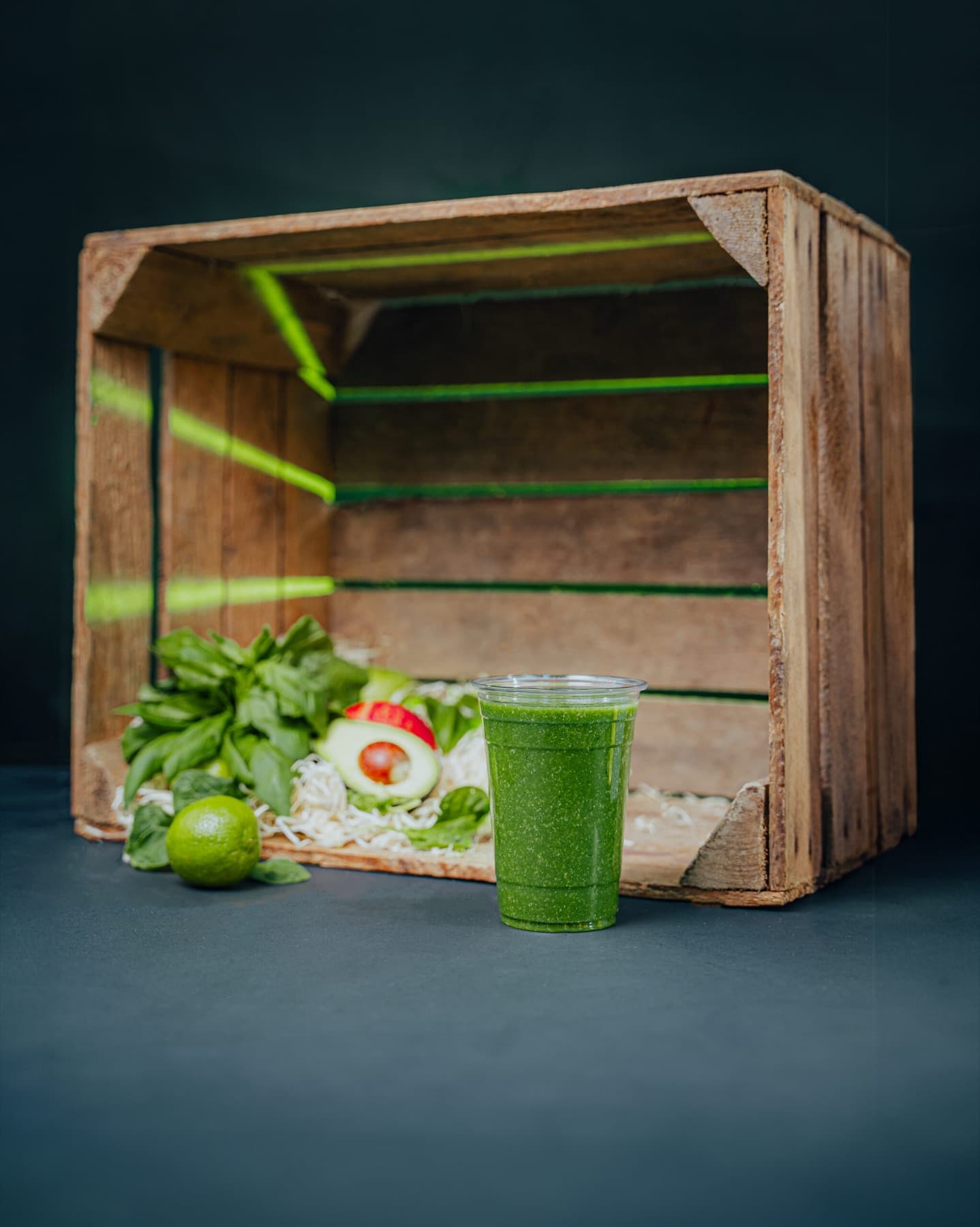A clear cup of vivid green Super Green smoothie on a dark studio set with greens around the base.