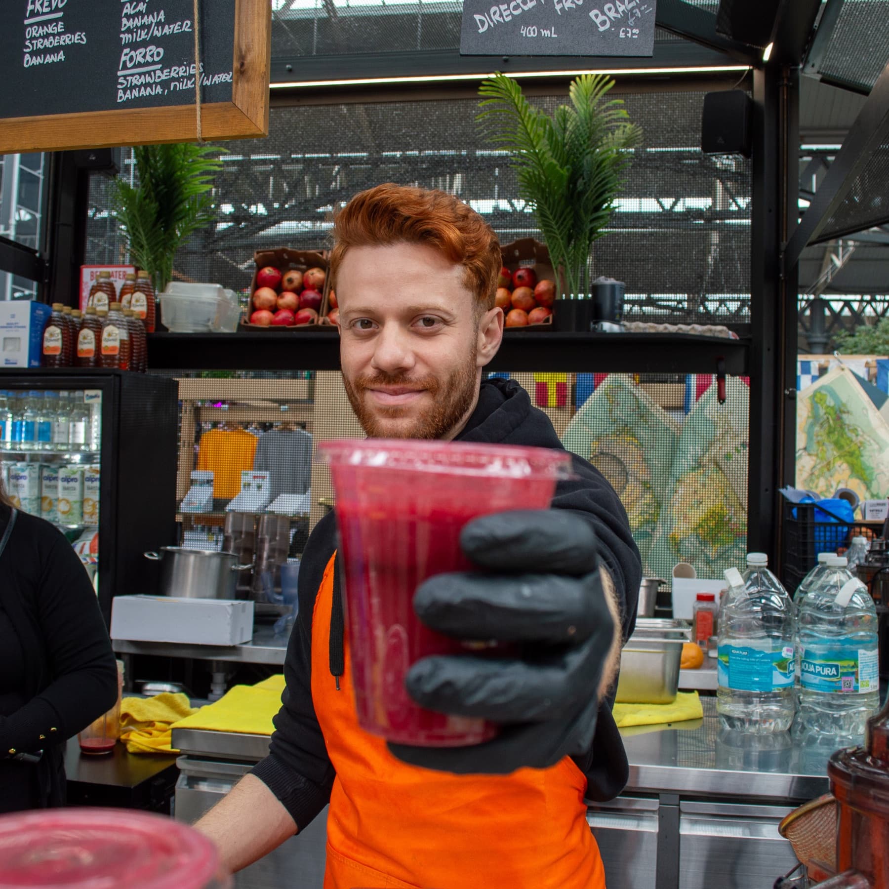 A Juice Guys team member in an orange apron handing over a freshly made juice at the market stall.