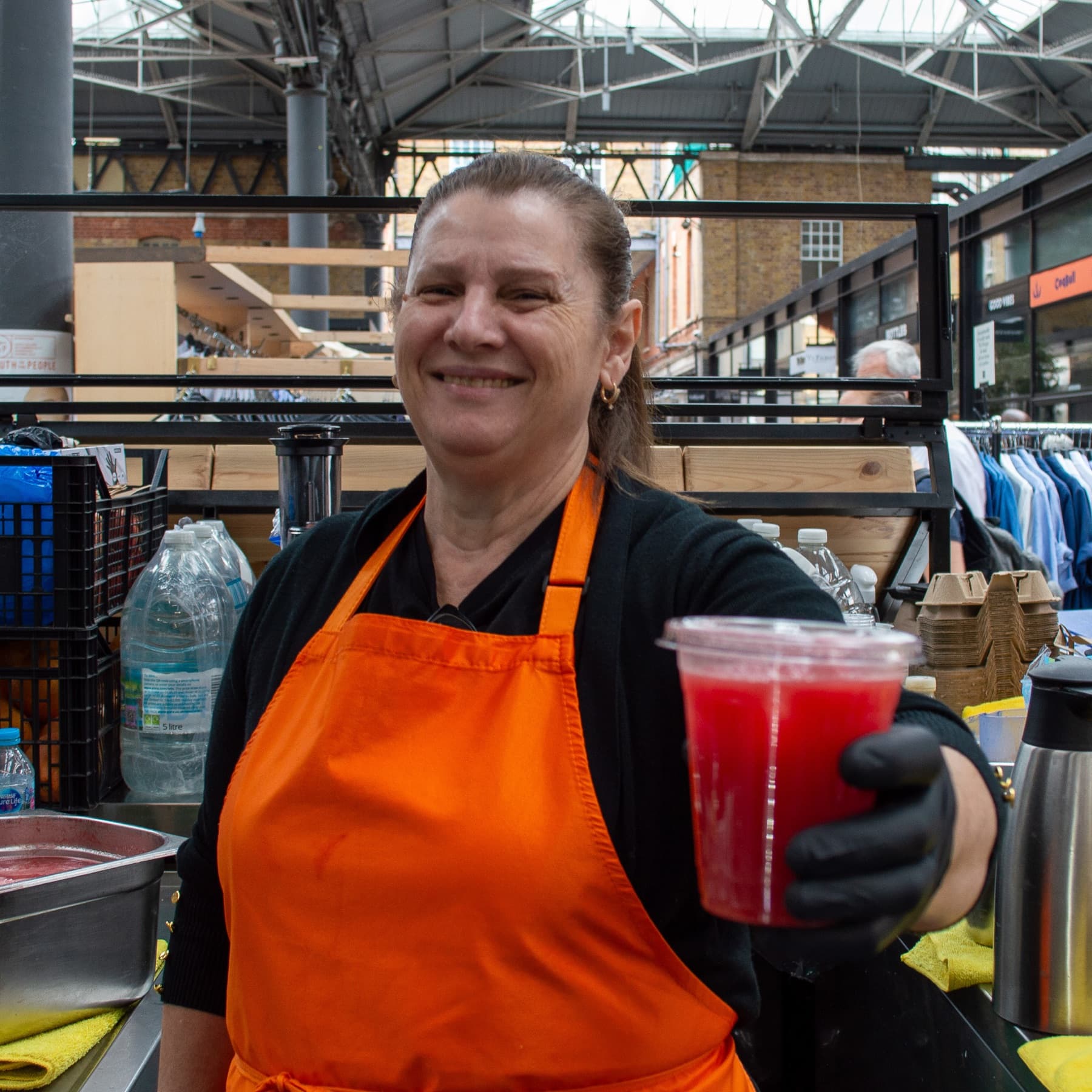 A smiling member of the Juice Guys family in an orange apron, handing over a fresh juice at the Old Spitalfields Market stall.